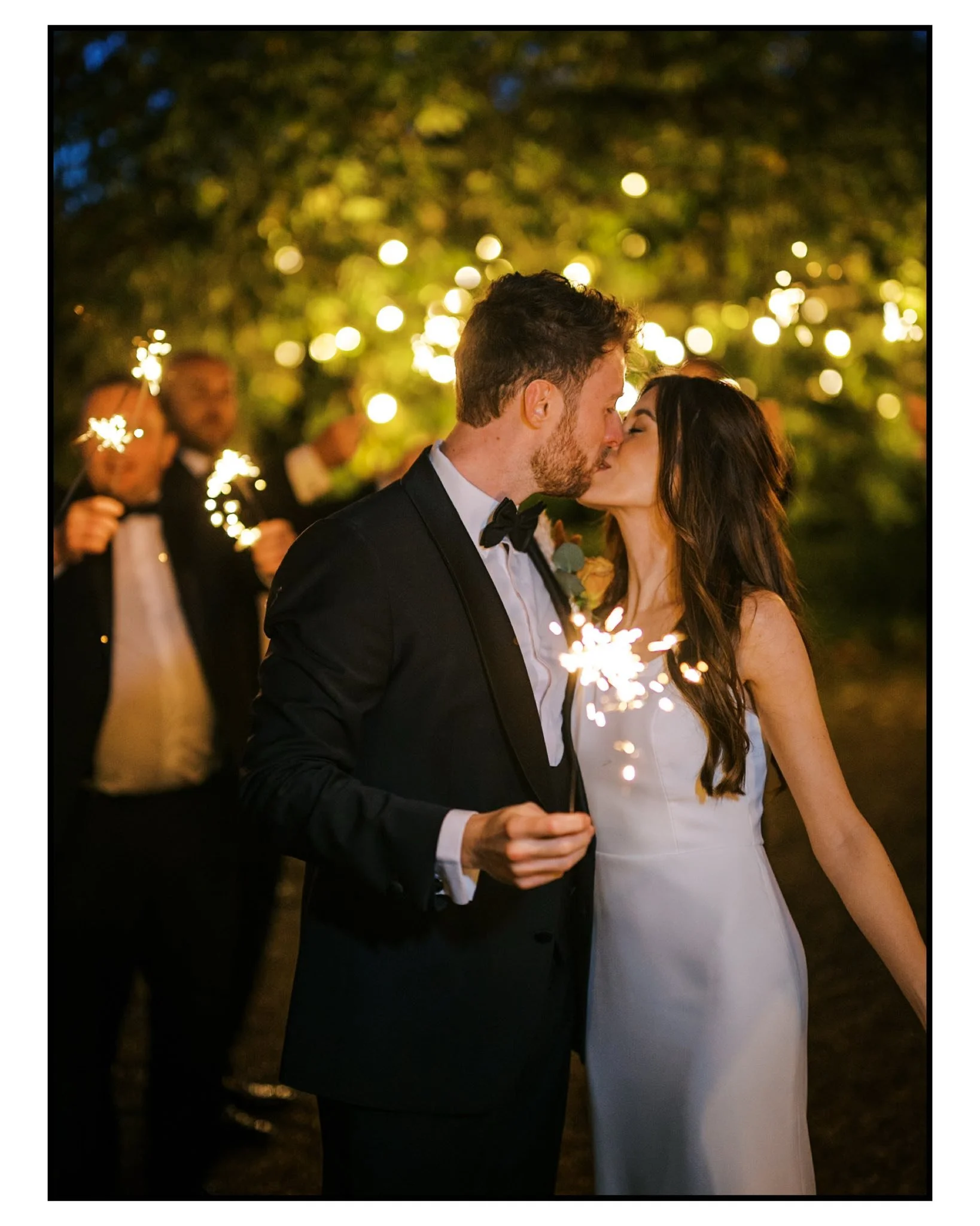 A newlywed couple in wedding attire sharing a kiss and holding sparklers during nighttime, with a background of trees and guests also holding sparklers. Take at Hodsock Priory