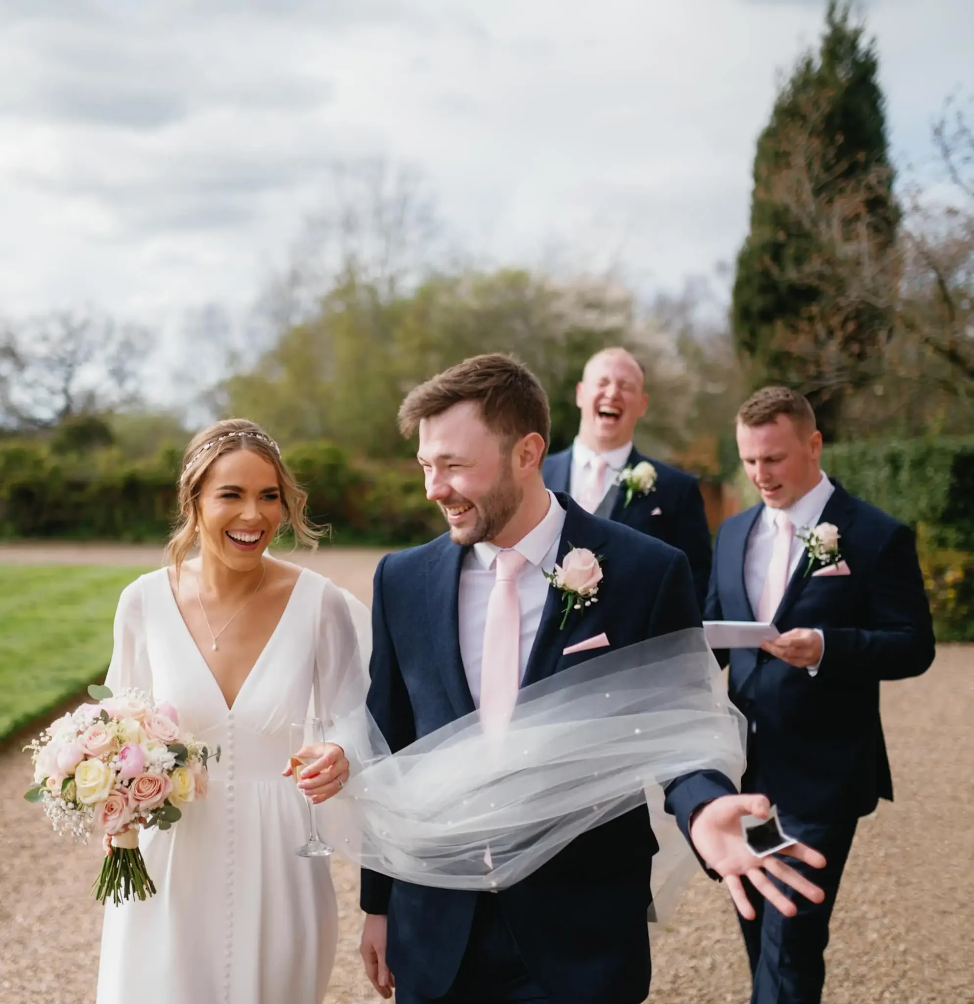 Group of four wedding guests walking outdoors on a cloudy day, smiling and laughing, with three men in navy suits and pink ties, and a woman in a white dress holding a bouquet, holding a smartphone. The background is the grounds of Hodsock Priory