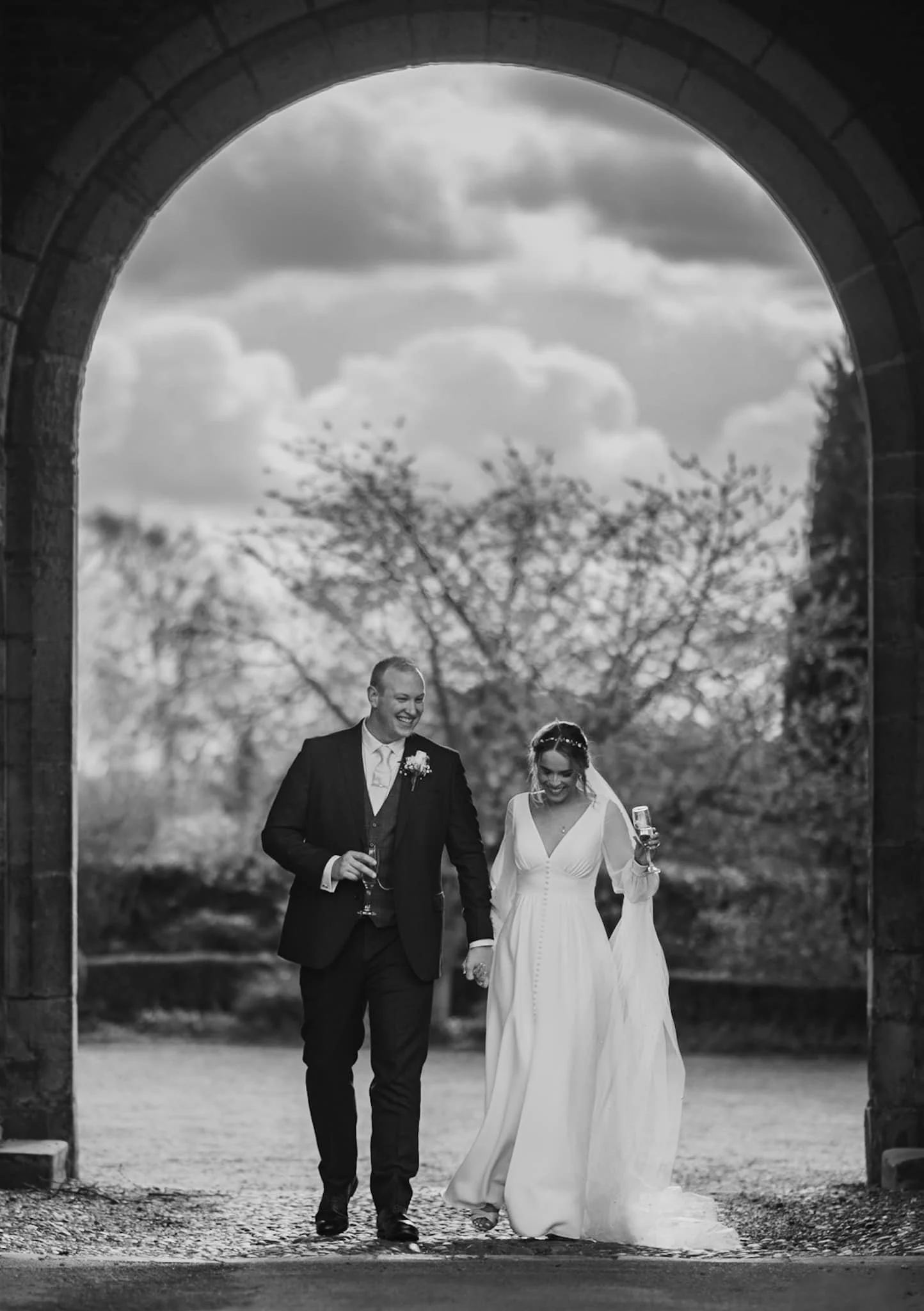 A black and white photo of a newly married couple walking hand-in-hand under an archway, holding champagne glasses and smiling. Taken at Hodsock Priory