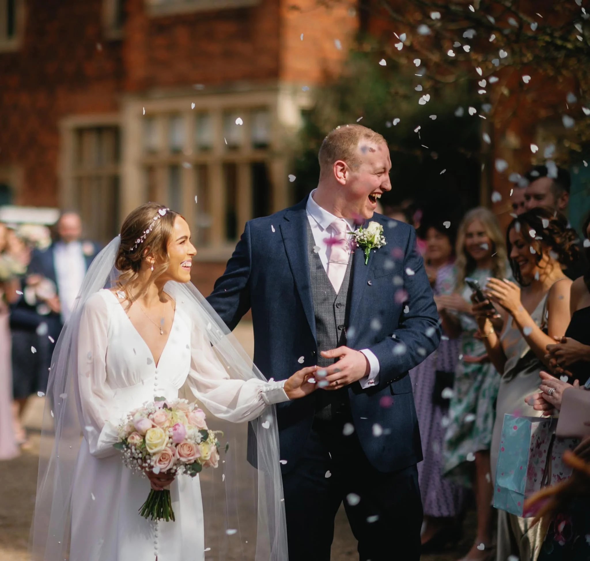 A bride and groom walking together after their wedding ceremony, smiling and celebrating among friends and family, with white confetti falling around them. Taken at Hodsock Priory