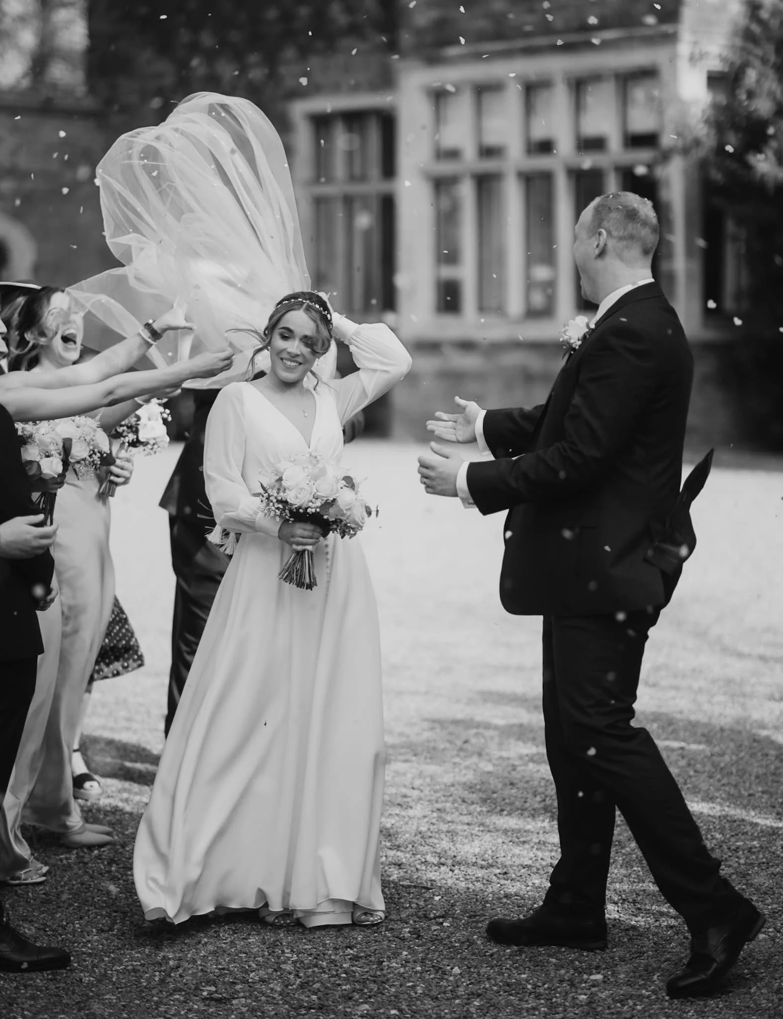 Black and white photo of a wedding scene outside, with a bride in a long dress holding a bouquet, smiling, and a groom in a suit, clapping. Guests are spreading confetti, and a woman is throwing a veil or fabric in the air. Take at Hodsock Priory