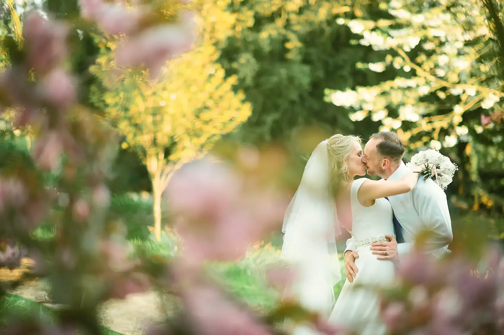 A couple in wedding attire sharing a kiss in a garden, surrounded by blurred pink and purple flowers and green foliage. Taken at Hodsock Priory