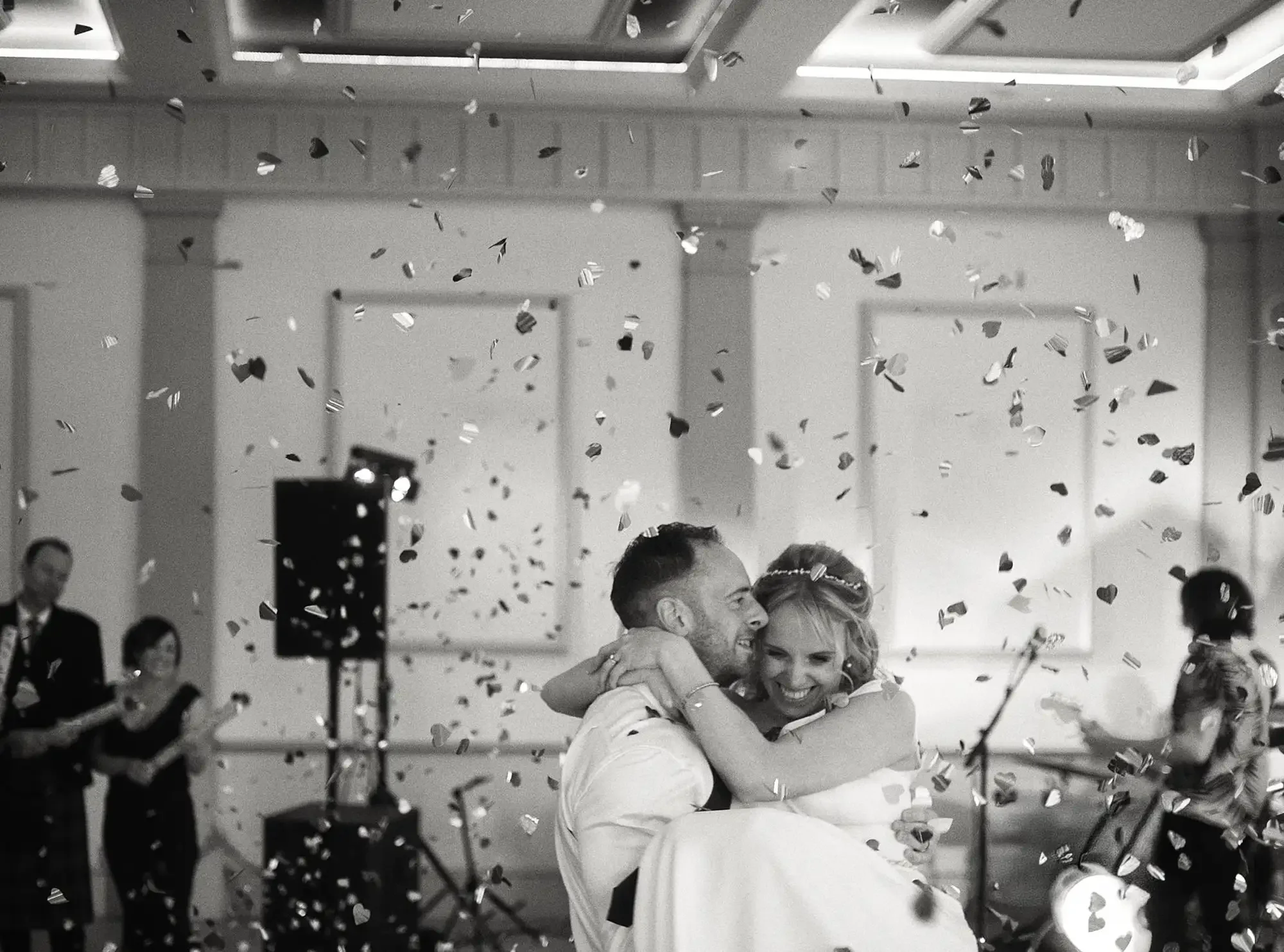 A bride and groom are dancing and hugging at their wedding reception, surrounded by falling confetti and smiling guests in the background. Taken at Hodsock Priory