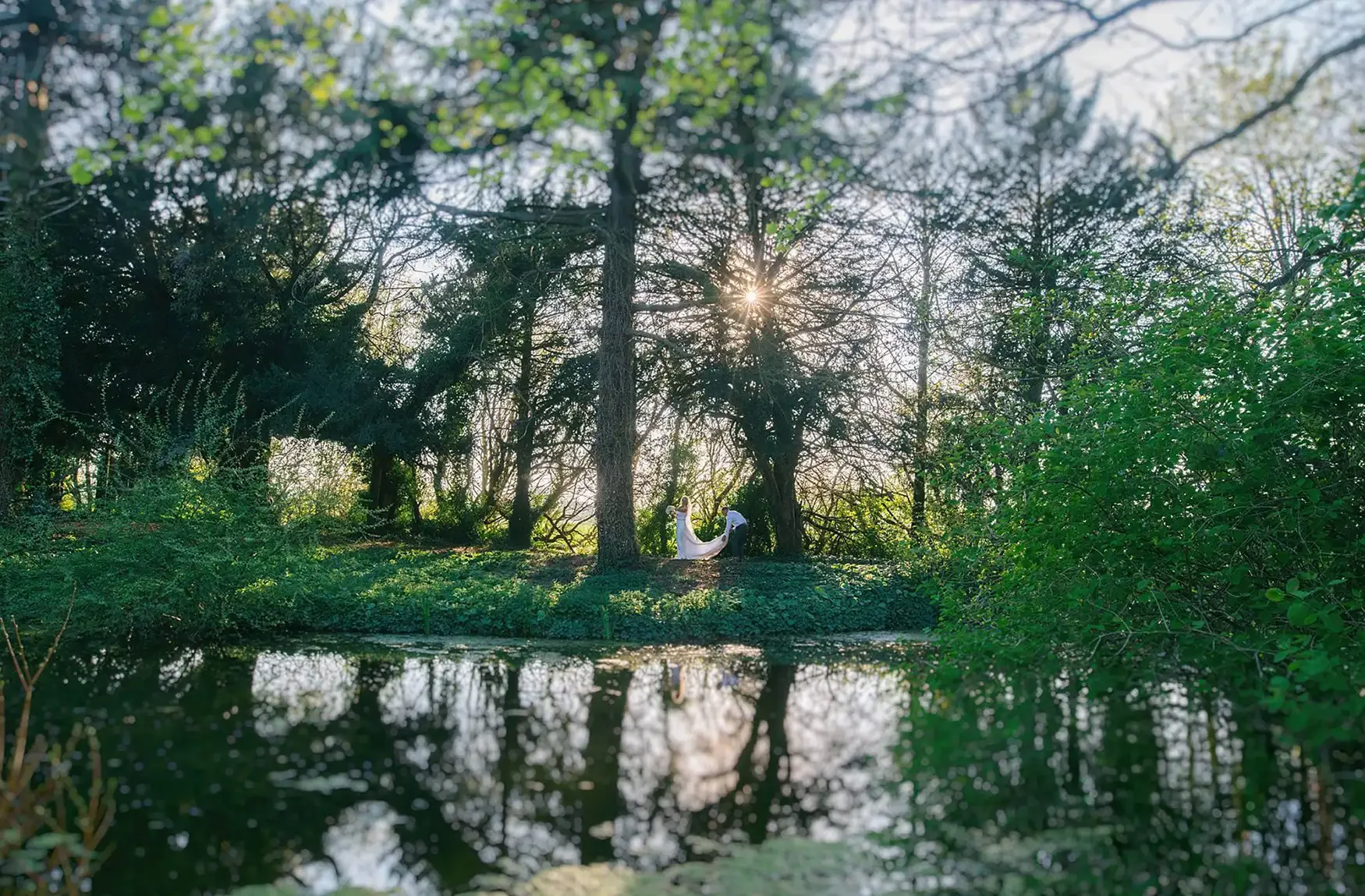 A bride and groom standing together by a large tree in a forested area during sunset, with a reflective body of water in the foreground. Take at Hodsock Priory
