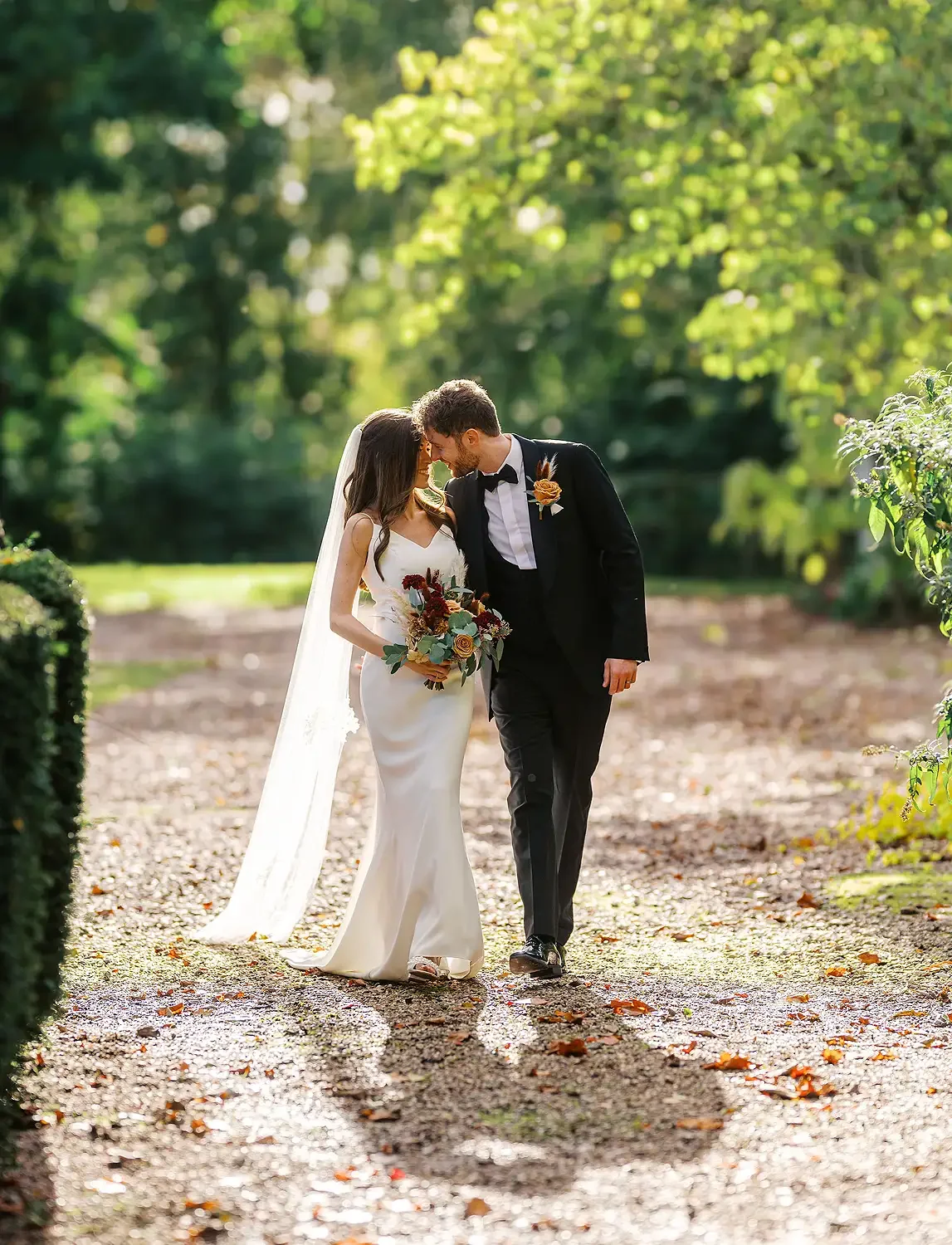 A bride and groom sharing a tender moment outdoors in a lush green park, with sunlight filtering through trees, the bride holding a bouquet of flowers, both leaning their foreheads together. Taken at Hodsock Priory