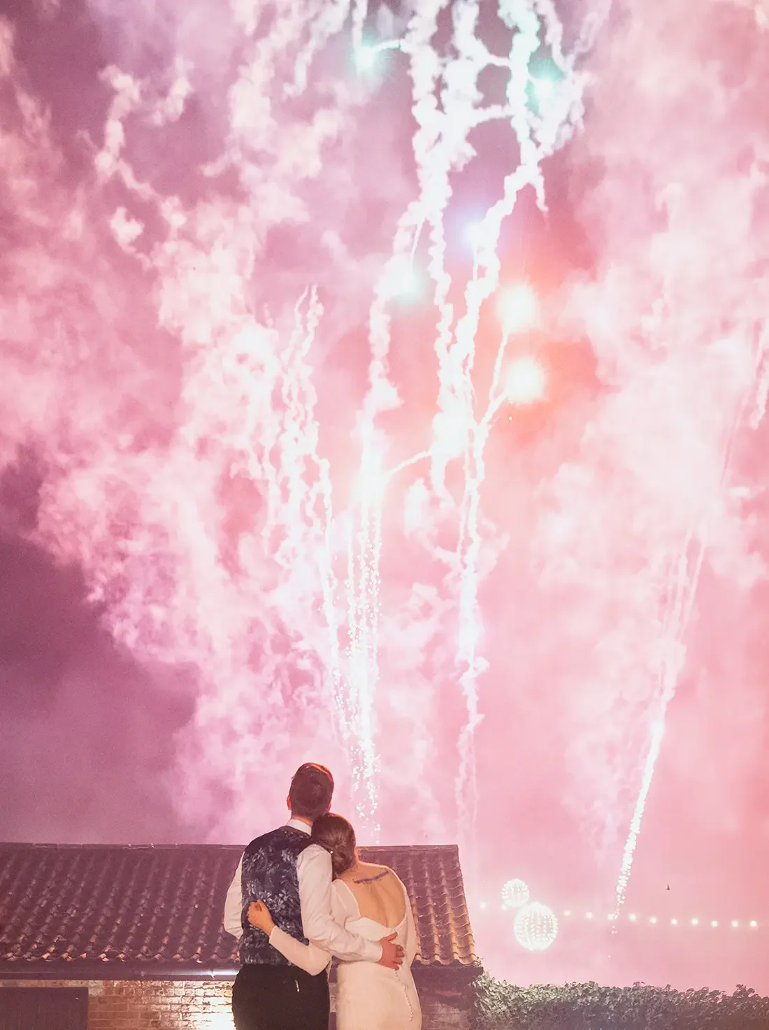 A couple dressed in formal attire, with the man in a dark vest and the woman in a white dress, stands hugging and watching fireworks in the night sky. The Normans wedding venue york