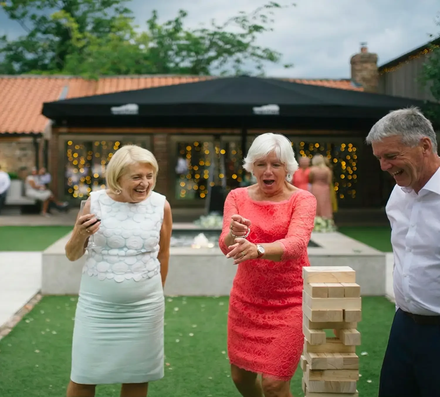 Three elderly adults playing a game of Jenga outdoors at a gathering, with a woman in a red dress pulling a block from the Jenga tower, two others watching and laughing, in the courtyard of the Normans wedding venue York