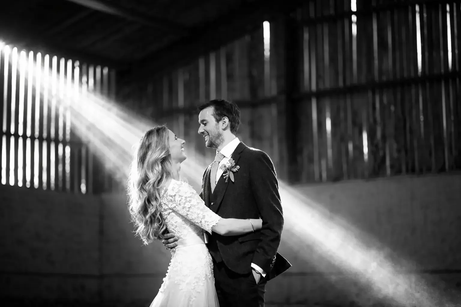 Black and white photo of a bride and groom sharing a dance, facing each other, in an indoor setting with wooden walls and sunlight streaming through gaps. Taken at The Normans wedding venue york