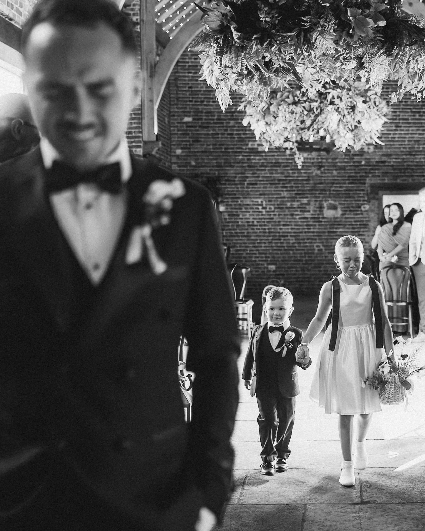 Black and white photo of a flower girl crying and a grinning page boy walking hand in hand down the aisle, with the groom welling up in the foreground