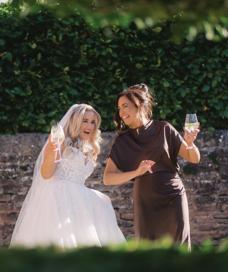 Bride and a friend laughing together with glasses of fizz in front of a hedge and old brick wall during the drinks reception