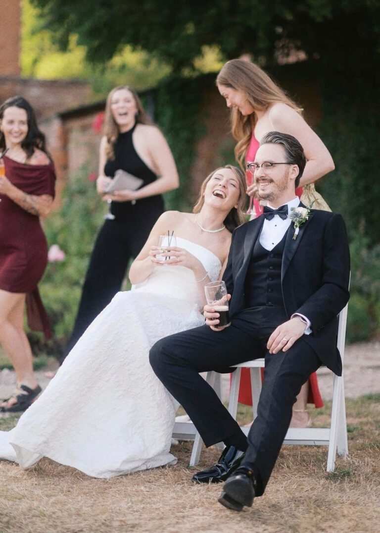 Bride slumped on a garden chair laughing, with the groom and bridesmaids laughing around her