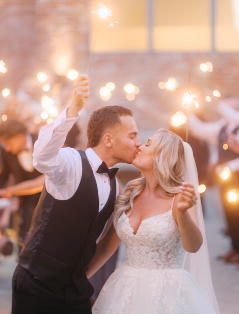 Groom in a waistcoat kissing the bride during a sparkler exit, guests holding glowing sparklers all around them